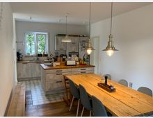 Helle Wohnküche mit modern-rustikalem Design, bestehend aus einer Küchenzeile mit grauen Holzfronten, einer zentralen Kücheninsel und einem langen Esstisch aus Holz mit grauen Stühlen. Über dem Tisch hängen drei schlichte, metallene Hängeleuchten, und ein Fenster neben der Küchenzeile bietet Blick ins Grüne.