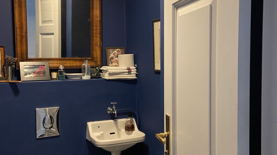 Kleines WC mit dunkelblauen Wänden, wandhängendem Waschbecken und Toilette sowie sechseckigen Bodenfliesen. Über dem Waschbecken hängt ein großer Spiegel mit Holzrahmen und einer schmalen Ablage mit Handtüchern und Pflegeartikeln; eine weiße Tür steht halb offen.
