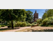 Ein historischer Backsteinturm mit spitzem Dach steht inmitten einer parkähnlichen Umgebung mit Bäumen, Sträuchern und Rasenflächen. Im Vordergrund führen sandige Wege durch den gepflegten Grünbereich, umgeben von dichten Hecken.