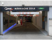 Einfahrt zu einem Parkhaus mit dem Schriftzug „Q-Park Märkische Zeile“. Über der Zufahrt befinden sich Schilder mit Höhen- und Geschwindigkeitsbegrenzung sowie ein LED-Display mit der Anzeige „Frei“.