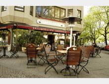 Außenbereich eines Restaurants mit dem Namen „Salumeria Via Lodovico“. Holztische und Stühle stehen auf einem gepflasterten Gehweg, umgeben von Pflanzen und Bäumen, mit einem Gebäude im Hintergrund und Heizstrahlern zwischen den Sitzbereichen.