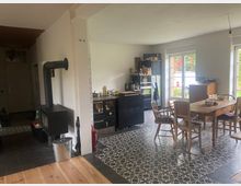 Ein Wohnraum mit offener Küche und Essbereich. Der Raum verfügt über gemusterte Bodenfliesen, einen schwarzen Holzofen, eine Küchenzeile mit schwarzem Mobiliar sowie einen Holztisch mit mehreren Stühlen. Tageslicht fällt durch große Fenster mit Blick auf einen Garten.