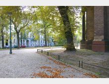 Kopfsteinpflasterstraße in einer baumgesäumten Umgebung mit einer historischen Backsteinfassade rechts im Bild. Im Hintergrund sind weiße Wohngebäude und parkende Autos zu sehen.