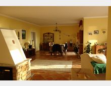 Wohnzimmer mit gelb gestrichenen Wänden, gefliestem Boden und einer offenen Gestaltung. Im Vordergrund ein gemauerter Kamin, im Hintergrund ein Esstisch mit Stühlen und Holzschränken; dekoriert mit Pflanzen und Wandbildern.