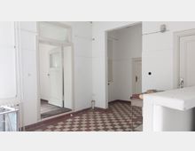 Ein einfacher Innenraum mit weißen Wänden, einer Tür mit Glaseinsatz sowie weiteren geschlossenen Türen. Der Boden ist mit rot-weiß karierten Fliesen ausgelegt, und an der rechten Seite ist ein Tresen oder Empfangstisch sichtbar.