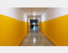 Ein langer, gelber Flur mit weißen Wänden im oberen Bereich, glänzendem Fliesenboden und mehreren Holztüren an beiden Seiten; am Ende des Flurs ist eine Glastür sichtbar.