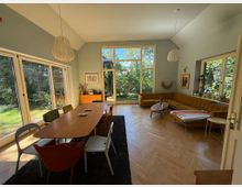 Helles Wohnzimmer mit großen bodentiefen Fenstern, die den Blick auf einen Garten freigeben. Im Raum befinden sich ein Holztisch mit bunten Stühlen, eine senffarbene Eckcouch, ein kleiner Couchtisch und helle Holzböden im Fischgrätmuster.