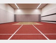 Ein Innenraum einer Sporthalle mit einer roten Sprintbahn, die in eine Sandgrube mündet, wie bei einer Weitsprunganlage. Die Wände sind grau und schlicht, die Decke mit integrierten Beleuchtungsstreifen versehen.