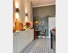 Helle Küche mit gemustertem Bodenfliesen in schwarz-weiß, einer weißen Schrankfront und einem dunklen, gemusterten Wandbild, das Bäume zeigt. Links an der Wand befinden sich drei goldene Wandleuchten, rechts ein kleiner runder Tisch mit Holzstuhl sowie eine Pendelleuchte mit gelbem Licht.