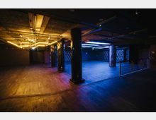 Großer, offener Veranstaltungsraum mit dunklem Holzboden, schwarzen Säulen und moderner Deckenbeleuchtung in Gelb- und Blautönen. Der Raum hat metallische Geländer und gemusterte Wände, die ein dezentes Ambiente schaffen.