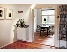 Innenraum einer Wohnung mit Holzboden. Im Vordergrund ein Flur mit einer Wanddekoration aus Bildern, einer Pflanze und Regalen; durch eine offene Tür ist ein Esszimmer mit einem Holztisch, vier Stühlen und einer hängenden Lampe zu sehen, dahinter ein Fenster mit Blick ins Freie.