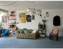 Eclectic living room with a patterned sofa, a blue upholstered chair, and a cane rocking chair on carpeted flooring. White walls are decorated with posters, hanging plants, and shelves holding glassware and small objects, with a homemade pendant lamp suspended from the ceiling.