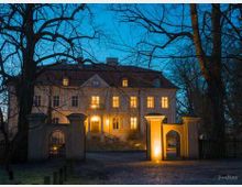 Herrenhaus in abendlicher Beleuchtung, mit mehreren erleuchteten Fenstern, umgeben von kahlen Bäumen. Im Vordergrund ein Torbogen mit angrenzendem Zaun, auf einem gepflasterten Weg.