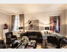 Wohnzimmer mit eleganter Einrichtung, schwarzem Polstermobiliar und dekorativen Elementen wie Kunstwerken an den Wänden, darunter ein Mickey-Mouse-Bild. Zentral steht ein Couchtisch mit Büchern und Blumen; Tageslicht fällt durch ein großes Fenster mit beigefarbenen Vorhängen.