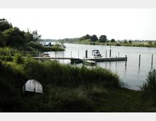 Eine Flusslandschaft mit einem kleinen Bootsanleger, an dem mehrere Boote angelegt sind. Im Vordergrund sind dichte Gräser und Büsche, während im Hintergrund Bäume die Uferlinie säumen.