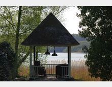 Ein Pavillon mit Spitzdach steht am Ufer eines Sees, umgeben von Bäumen und Schilf. Im Inneren des Pavillons befinden sich Sitzmöbel und zwei hängende Lampen.