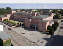 Luftaufnahme eines weitläufigen, rosa verputzten Gebäudekomplexes im Bauhausstil mit rechteckigen Fenstern und flachen Dächern. Der Komplex ist von einem gepflasterten Platz mit Parkplätzen, Grünflächen und Hecken umgeben.