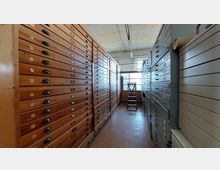 Ein schmaler Archivraum mit hohen Holz- und Metallschränken, die aus vielen übereinander angeordneten flachen Schubladen bestehen. Im Hintergrund steht eine kleine Leiter vor einem Fenster mit mattierter Verglasung.