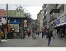 Straßenszene am Theodor-Heuss-Platz mit einem U-Bahn-Eingang, der durch ein blaues Schild gekennzeichnet ist. Im Vordergrund Gehwege mit Fußgängern und Fahrrädern, flankiert von Geschäftsgebäuden mit Schaufenstern und Cafés; im Hintergrund sind Bäume und mehrstöckige Gebäude sichtbar.