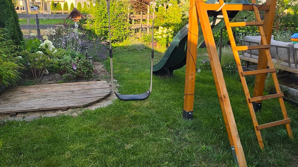 Garten mit Rasenfläche und Holz-Spielgerüst mit Schaukel und Leiter; dahinter stehen Beete mit Sträuchern und Bäumen. Im Hintergrund sind Wohnhäuser und ein blauer Himmel mit Wolken zu sehen.