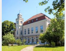 Ein hellbeiges, zweigeschossiges Gebäude mit roten Dachziegeln, großen Fenstern und einem markanten Turm an der linken Seite. Es ist umgeben von Bäumen, Büschen und einer gepflegten Grünfläche unter einem klaren, blauen Himmel.