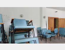 Ein leerer Raum mit gestapelten, blauen Stühlen mit Metallrahmen. Im Hintergrund sind weiße Wände, zwei hölzerne Türen und Deckenleuchten sichtbar.