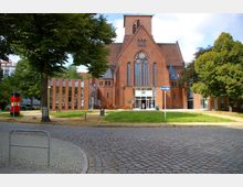 Backstein-Kirchengebäude mit großen gotischen Fenstern und einem modernen Eingangsbereich aus Glas, umgeben von Bäumen und einem gepflasterten Platz im Vordergrund. Links ist ein Nebengebäude mit vertikalen Glas- und Backsteinelementen zu sehen.