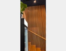 Holzverkleidete Wand aus vertikalen Holzlatten, die eine Treppe mit passendem Holzgeländer flankiert, rechts ein schwarzer Deckenbereich; links ein beleuchteter Kühlschrank und eine große grüne Pflanze.