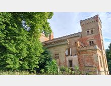 Eine verfallene Backsteinvilla mit Zinnen und einem Turm, umgeben von dichter Vegetation und Bäumen. Die Fassade ist teilweise beschädigt und überwuchert, Fensteröffnungen sind teilweise nicht mehr verglast.