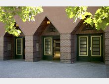 Eingangsbereich eines Gebäudes mit drei grünen Holztüren, umgeben von gemauerten, braun-roten Backsteinbögen. Darüber befinden sich dreieckige Verzierungen und ein schlichtes Putzfassadendach; Vordergrund mit gepflastertem Boden und Blättern eines Baumes.