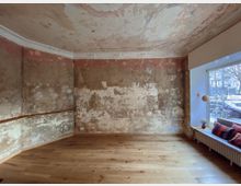 Ein minimalistisch eingerichteter Raum mit verwitterten, scheinbar unrenovierten Wänden und einer Decke mit verblassten Verzierungen. Der Boden besteht aus hellem Holz, ein großes Fenster rechts lässt Tageslicht herein, und davor steht eine Sitzbank mit bunten Kissen.