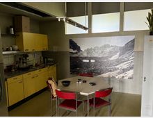 Blick in eine Gemeinschaftsküche mit gelben Schrankfronten, Edelstahl-Arbeitsbereich und einem sechseckigen Tisch mit roten Stühlen. An der Wand hängt ein großformatiges Schwarzweiß-Foto einer Berglandschaft, darüber befinden sich hohe Fenster.