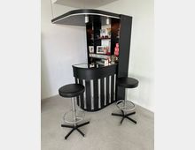 Schwarze Hausbar mit geschwungenem Dach und integrierter Beleuchtung, darunter Regale mit Gläsern, Dekorationen und Getränken; davor zwei schwarze Barhocker mit Chromfüßen auf hellgrauem Boden.