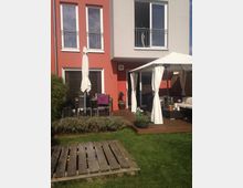 Wohnhaus mit moderner Fassade in Rot und Weiß, ergänzt durch einen Garten mit Rasenfläche. Im Vordergrund eine Holzterrasse mit Gartenmöbeln, einem Pavillon mit weißen Vorhängen und Pflanzenarrangements.