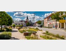 Kleinstadtzentrum mit einer gepflasterten Fußgängerzone, umgeben von Bäumen, Blumenbeeten und Sitzbänken im Vordergrund. Im Hintergrund sind Geschäfte und ein Kirchturm mit spitzem Dach zu sehen, während ein blauer Himmel mit weißen Wolken die Szene umrahmt.
