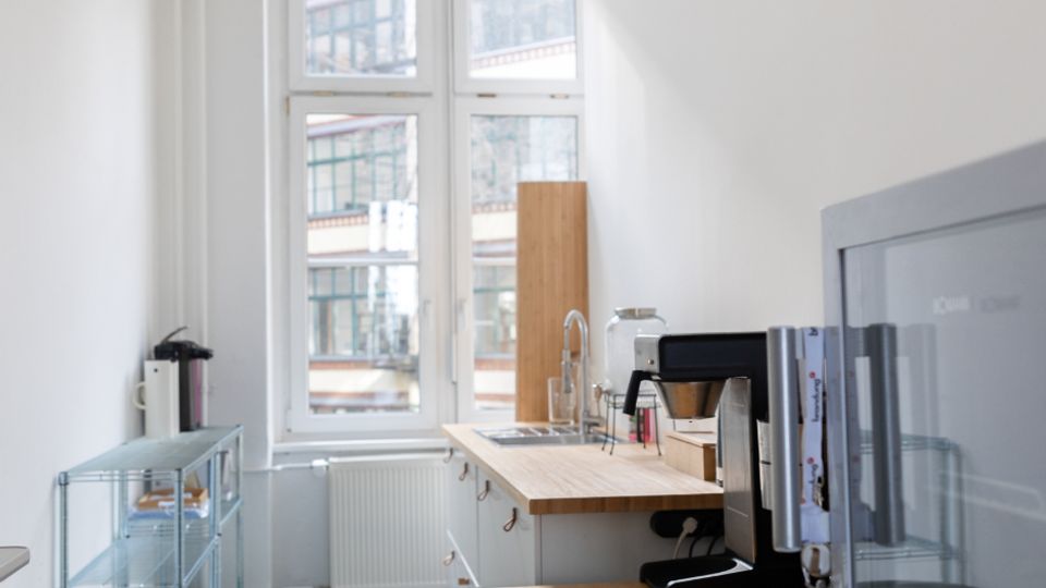 Schmale, helle Büroküche mit großen Sprossenfenstern, grauem Boden und weißen Wänden. Entlang der rechten Seite stehen Arbeitsplatten aus Holz mit Spüle und Kaffeemaschine, links ein Metallregal und ein Abfalleimer.