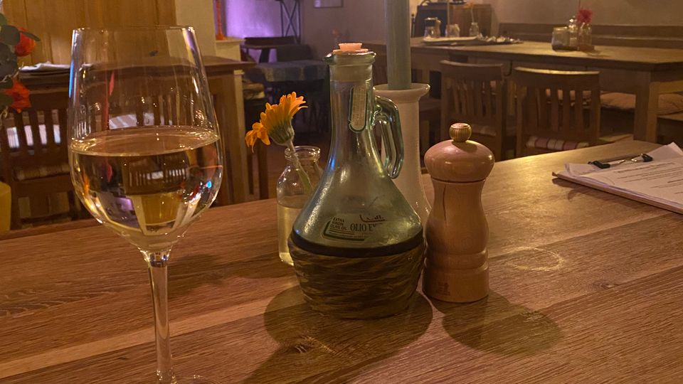 Innenraum eines gemütlichen Restaurants mit Holztischen und -stühlen, warmen Wandlampen und Bildern an den Wänden. Im Vordergrund steht auf einem Holztisch ein Weinglas sowie Kerze, Glasflasche, kleine Blumenvase und Gewürzmühle.