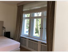 Ein helles Schlafzimmer mit großem, weißem Sprossenfenster, umgeben von beige Vorhängen. Unter dem Fenster befindet sich ein Heizungsgitter, und im Raum steht ein Bett mit heller Bettwäsche.