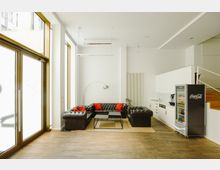 Moderner Aufenthaltsraum mit einer braunen Ledercouch in L-Form, roten Kissen, einem schwarzen Couchtisch und einer Stehlampe. Im Hintergrund befinden sich eine kleine Küchenzeile, ein Getränkeautomat und eine Treppe, die nach oben führt. Große Glastüren auf der linken Seite lassen Tageslicht herein.