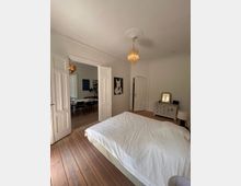 Ein helles Schlafzimmer mit Holzboden und einem großen Bett mit weißer Bettdecke. Links führen weiße Doppeltüren in einen angrenzenden Raum mit einem Esstisch und Stühlen; rechts stehen eine Kommode mit Spiegel und eine Tür. An der Decke hängt ein goldfarbener Kronleuchter.