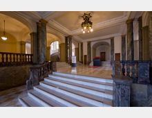 Eingangshalle eines historischen Gebäudes mit Marmorsäulen, weißem Treppenaufgang und dunklen Balustraden. Im Hintergrund befinden sich dekorative Bögen, Holztüren und ein Kronleuchter an der Decke.
