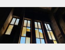 Ansicht eines verlassenen, heruntergekommenen Gebäudes mit hohen, beschädigten Fenstern, durch die teils zerrissene Glasscheiben den Blick auf den blauen Himmel freigeben. Die Wände bestehen aus rotem Backstein und Beton und vermitteln eine industrielle Atmosphäre.