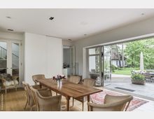 Helles Esszimmer mit einem Holztisch und sechs Korbstühlen, bodentiefen Fensterfronten zur Terrasse und Blick in einen grünen Garten mit Rasen, Bäumen und einem kleinen Haus im Hintergrund. Links führen Glastüren zu einem Treppenhaus, und ein Teil eines roten Teppichs ist auf dem Holzboden sichtbar.