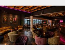 Innenraum einer modernen Lounge-Bar mit vielen runden Tischen und gepolsterten Samtsesseln in verschiedenen Farben, auf einem glänzenden Boden. Dunkle Wände mit Kunstbildern, große Fensterfronten, Säulen sowie eine kassettierte Decke mit warmem Licht und violetten LED-Akzenten prägen den Raum.
