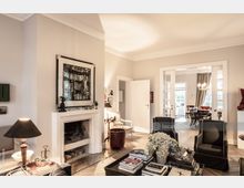 Helles Wohnzimmer mit hellbeigen Wänden, einem Kamin, darauf ein gerahmtes Schwarz-Weiß-Bild, sowie einer Einrichtung mit modernen Möbeln. Im Hintergrund ist ein Essbereich mit einem Kronleuchter und großen Fenstern zu sehen.