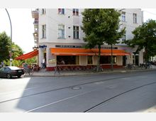 Eine Straßenecke mit einem indischen Restaurant und Café mit der Aufschrift "Indisches Restaurant-Café-Cocktailbar", erkennbar an den orangen Markisen. Vor dem Gebäude stehen Fahrräder, ein Auto und kleine Tische im Außenbereich, umgeben von Bäumen und Tramgleisen im Vordergrund.
