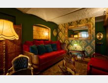 Gemütliches Wohnzimmer im Vintage-Stil mit grünen Wänden, gold-verzierter Tapete und antikem Holzmobiliar. Zu sehen sind ein rotes Sofa mit farbigen Kissen, eine große Stehlampe, ein hölzernes Schaukelpferd und ein großer goldgerahmter Spiegel an der Wand.