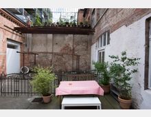 Hinterhof mit Backstein- und verputzten Wänden, einem Tisch mit rot-weiß kariertem Tischtuch, umgeben von Pflanztöpfen. Darüber befindet sich ein Metallbalkon mit weiteren Pflanzen in Töpfen.