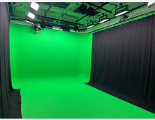 Ein professionelles Film- oder Fotostudio mit einem grünen Hintergrund (Greenscreen) und schwarzen Vorhängen an der Seite. An der Decke befinden sich mehrere Scheinwerfer und Studiolichter.