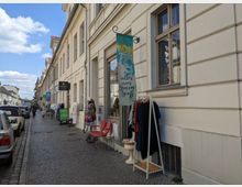 Schmale Kopfsteinpflasterstraße mit hellen, mehrstöckigen Altbauten; im Vordergrund ein Schaufenster mit Kleiderständer, daneben ein Schild mit dem Text „alles aus gutem Stoff“. Autos sind am Straßenrand geparkt und Passanten laufen entlang der Geschäfte.